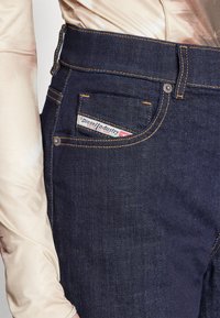 Dark denim jeans with golden stitching, high waist, and branded tag visible in front pocket. Smooth texture and classic five-pocket design.