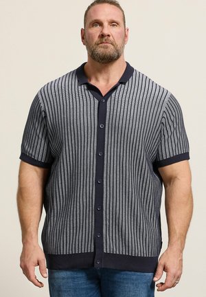 Homme barbu portant une chemise à manches courtes, boutonnée, rayée verticalement bleu marine et grise, et un jean bleu, debout devant un fond uni.