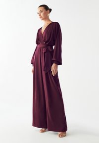 Burgundska zavita kombinezon s dolgimi rokavi in zavezanimi pasom. Ima globok V-izrez in leteči širok kroj nog. Teksturna tkanina z gladko površino.
