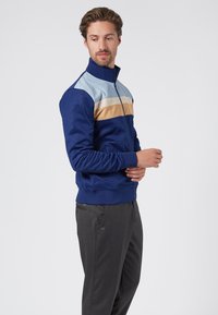 Navy zip-up jacket with light blue, beige, and white horizontal stripes, featuring a collar and ribbed cuffs, paired with dark gray trousers.