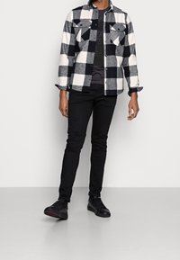 Schwarz-weiß kariertes Flanellhemd mit zwei Brusttaschen, über einem dunklen T-Shirt getragen, kombiniert mit schwarzen Slim-Fit-Jeans und schwarzen Turnschuhen.