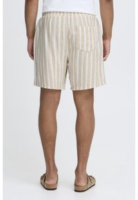 Shorts en coton beige et blanc à rayures avec une taille élastique et une poche arrière unique, portés avec des sandales marron.
