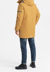 Parka amarilla con acentos grises reflectantes, combinada con jeans azul oscuro y botas negras, con capucha y diseño hasta la rodilla.