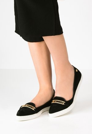 Nainen pukeutuneena mustiin slip-on-kenkiin, joissa on kultainen "Love Moschino" -logo ja valkoiset pohjat, yhdistetty mustiin lyhennettyihin housuihin.