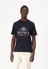 Tums zilgans kokvilnas krekls ar baltu izdrukātu tekstu "LES DEUX Sporting Goods", dekoratīviem elementiem, apaļu kakla izgriezumu un īsām piedurknēm.