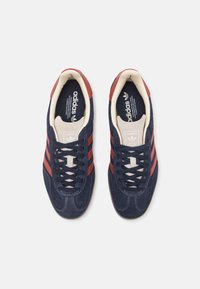 Blå mockasinliknande sneakers med röda accentremsor, krämfärgade skosnören, rundad tå och texturerad yta, med adidas-logotyp på tungan.