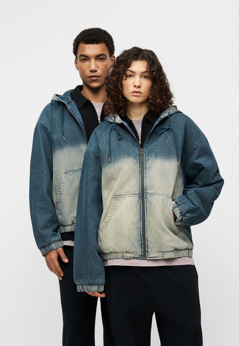 Τζιν μπουφάν με διαβάθμιση από σκούρο σε ανοιχτό μπλε, με μεγάλο μέγεθος, κουκούλα με κορδόνι και ελαστικές μανσέτες, φορεμένο από δύο μοντέλα.