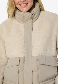 Uśmiechnięta kobieta w beżowym polarze sherpa i pikowanej kurtce z wysokim kołnierzem oraz dużymi przednimi kieszeniami, skróconej do ramion i ust.
