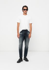 Hombre con gafas de sol negras, camiseta blanca de manga corta, jeans negros desgastados, calcetines negros y mocasines negros de pie contra un fondo blanco.