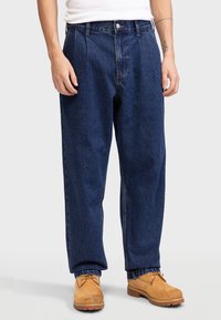 Pantalon en denim bleu foncé avec une coupe décontractée, devant plissé et coutures contrastantes. Porté avec des bottes beiges.