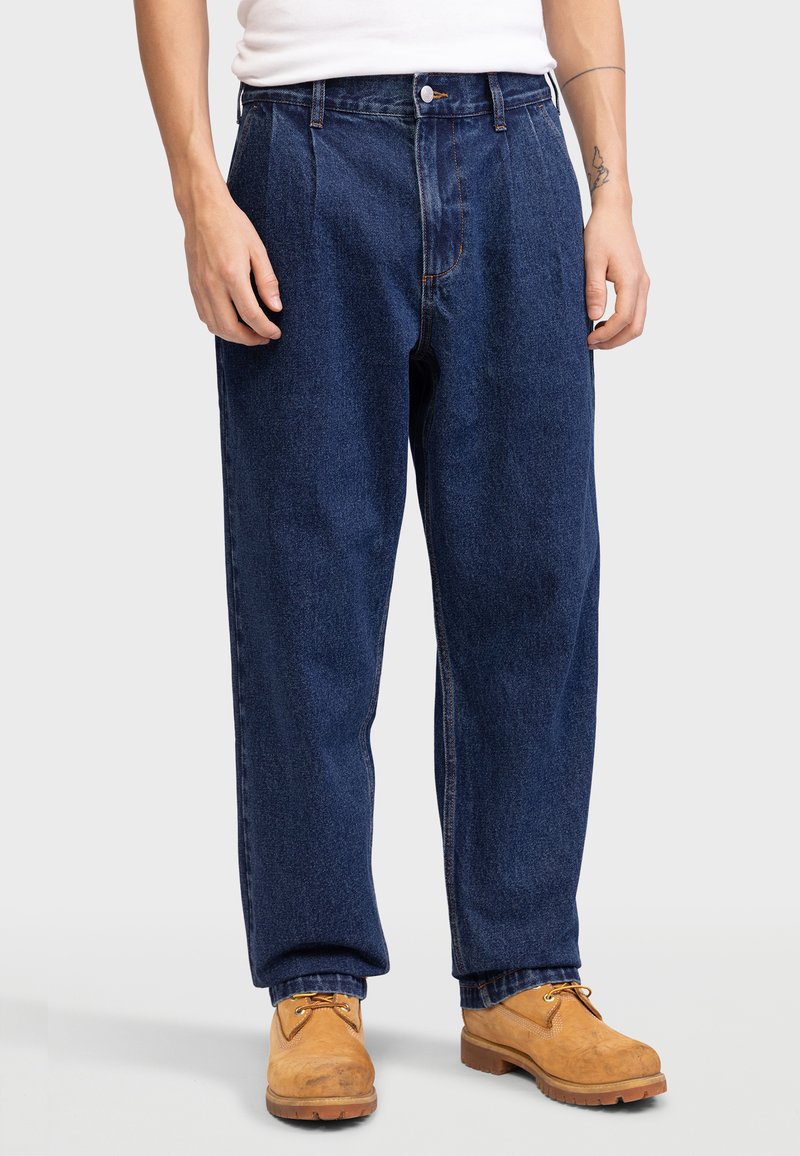 Pantalon en denim bleu foncé avec une coupe décontractée, devant plissé et coutures contrastantes. Porté avec des bottes beiges.