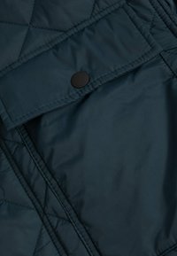 Ciemnoniebieska, pikowana tkanina z przeszytymi skośnymi liniami i czarnym zatrzaskiem na klapce kieszeni.