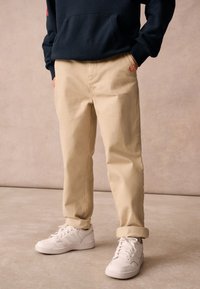 Pantaloni beige con vestibilità rilassata, tessuto leggero, tasche laterali e un'etichetta rossa sulla vita. Abbinati a sneaker bianche.