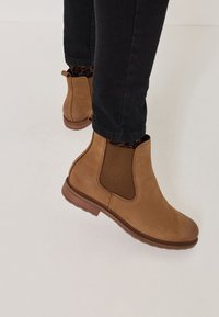 Persona che indossa stivali Chelsea in suede beige con pannelli laterali elastici neri, calzini con stampa leopardata e pantaloni neri, in piedi su un pavimento chiaro.