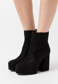 Bottes noires en daim à cheville avec un talon haut épais, un bout arrondi et des détails de coutures latérales. Texture lisse avec un éclat subtil.