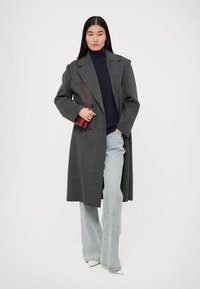 Cappotto di lana grigio con ampi revers, bottoni doppi e tasche frontali, indossato su un dolcevita blu navy e jeans a zampa di elefante blu chiaro.