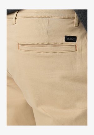 Beige katoenen broeken met een platte achterzak met een zwart label met de tekst "NINETEEN NINETY THREE" en oranje stikselaccenten.