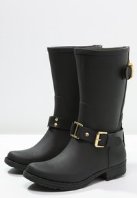 Black rubber rain boots with a rounded toe, mid-calf height, accentuated with gold-tone buckles and hardware, featuring a textured sole.