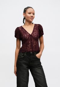 Blusa de renda borgonha com mangas curtas bufantes, decote em V e fecho frontal com botões. Combinada com jeans pretos de cintura alta. Padrão floral texturizado.