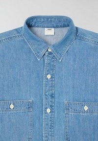 Lys blå denimskjorte med spredt krave, to brystlommer og knaplukning. Har et tekstureret stof og hvide plastikknapper.