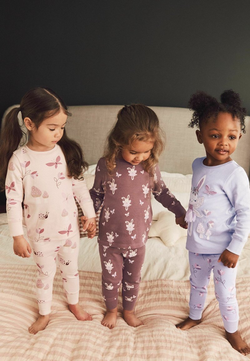Tre unge piger i mønstrede pyjamas holder hinanden i hånden og står barfodede på en seng med en beige gavl og en mørk væg bagved.