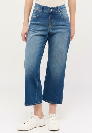 Blaue Denim-Jeans mit weitem Bein und hoher Taille, ausgestattet mit fünf Taschen, leichter Used-Optik und rohem Saum, kombiniert mit weißen Sneakern.