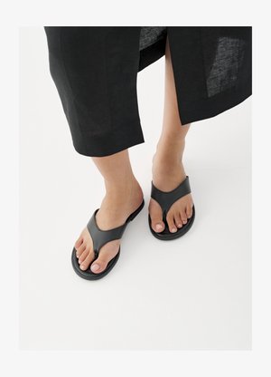 Jalutuskäik mustade nahast varbavahedega sandaalidega, musta seeliku ääreosa paistmas pahkluude kohal valgel põrandal.