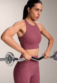 Donna con top sportivo color malva e leggings che solleva un bilanciere con pesi, concentrata sull'allenamento della forza su uno sfondo neutro.