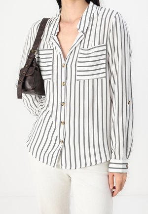 Femme portant une chemise blanche à manches longues avec des rayures verticales noires et des poches avant, portant un petit sac à bandoulière en cuir marron foncé.