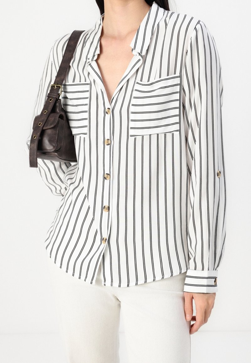 Femme portant une chemise blanche à manches longues avec des rayures verticales noires et des poches avant, portant un petit sac à bandoulière en cuir marron foncé.
