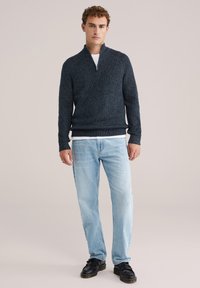 Tamborēta tumši zila zipa džemperis ar strukturētu rakstu, apvienots ar gaiši ziliem džinsiem un melnām kurpēm. Modelis stāv pret neitrālu fonu.