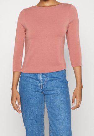 Femme portant un haut ajusté à manches longues rose poudré et un jean bleu taille haute, debout devant un fond clair uni.