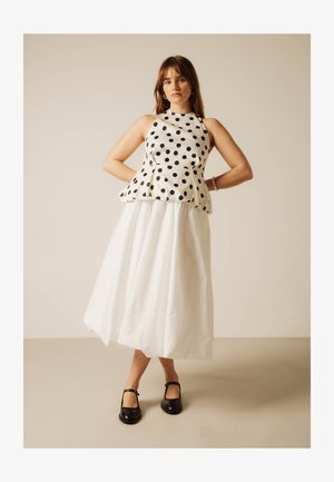 Femme debout, mains sur les hanches, portant un haut blanc sans manches à pois noirs, une jupe midi blanche et des chaussures Mary Jane noires.