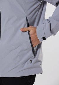 Veste grise à rayures verticales avec une poche zippée sur le côté, des poignets ajustables et des accents noirs, fabriquée à partir d'un tissu lisse et léger.