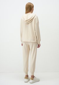 Beige hoodie met een rugventilatie en trekkoord, gecombineerd met bijpassende beige joggers met elastische boorden, gedragen met witte sneakers.