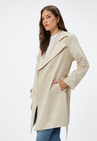 Cappotto trench beige doppiopetto con rever ampi, tasche laterali e dettagli in bottoni, realizzato in tessuto morbido con vestibilità su misura.