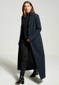 Cappotto lungo in misto lana blu navy con rever a punta, chiusura a singolo bottone e tasche laterali; abbinato a un dolcevita nero e jeans skinny.