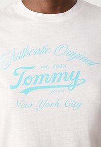Hvit bomull t-skjorte med turkise vintage-inspirerte bokstaver: "Authentic Original Tommy jeans New York City" med året "est. 1985."