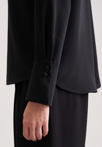 Blusa nera a maniche lunghe con una texture liscia. Il polsino presenta due bottoni rotondi e un orlo leggermente curvo. Design semplice e sartoriale.