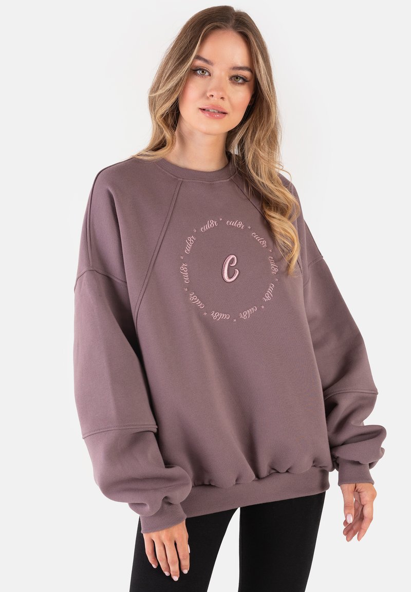 Przesadny lawendowy sweatshirt z ribbowanymi mankietami i dołem, z haftowanym okrągłym napisem oraz centralnym wzorem litery "C". Gładka faktura.