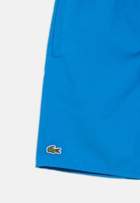 Kék fürdőnadrág könnyű anyagból, alsó szegélyén logó folttal. Az anyag oldalán zsebek és sima tapintású textúra található.