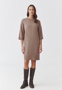 Robe en maille marron à manches courtes, coupe droite et ourlet côtelé. Associée à des bottes en cuir marron foncé jusqu'aux genoux.