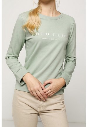 Femme portant une chemise à manches longues vert clair avec le texte « POLO CLUB Established 2012 », associée à un pantalon beige, mains jointes devant.
