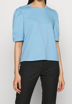 Kvinde iført en lyseblå top med korte ærmer og puf-ærmer og sorte bukser, stående mod en ensfarvet baggrund.