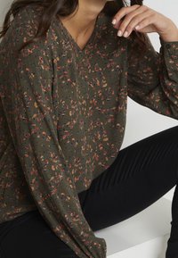 Blouse à manches longues vert olive avec un col en V, présentant un motif floral rouge, jaune et violet sur un tissu léger texturé.