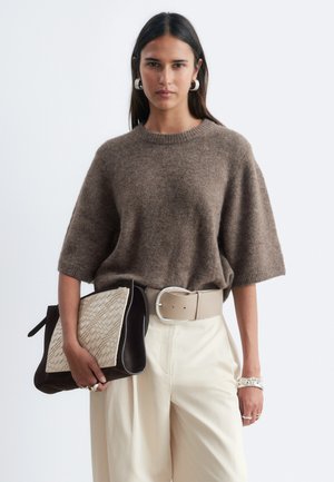 Femme portant un pull marron à manches courtes et un pantalon large crème tient une grande pochette tissée bicolore avec des boucles d'oreilles anneaux en argent.