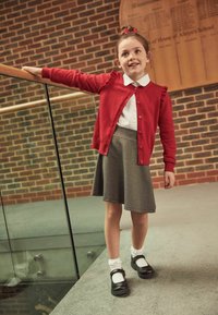 Red cardigan with ruffle detail, white button-up shirt, gray skirt, and black Mary Jane shoes with white ankle socks. Smooth, casual materials.