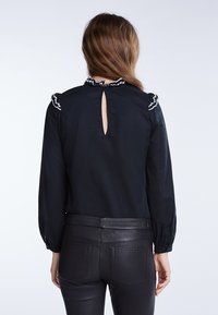 Sort bluse med lange ærmer, struktureret stof, flæsekrave med kontrasttrim og en nøglehulåbning bagpå. Kombineret med sorte bukser.