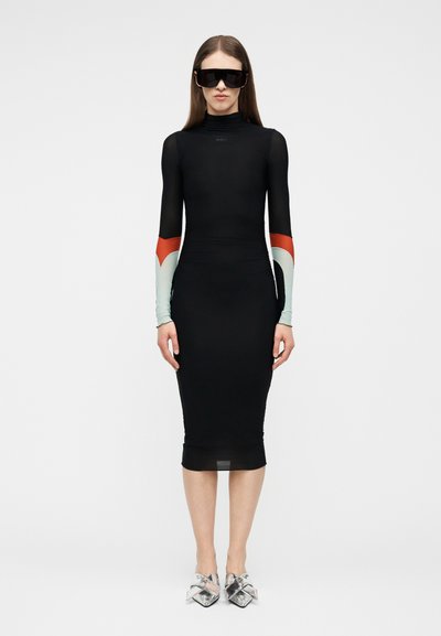 Nő, aki testhezálló fekete midi ruhát visel hosszú ujjakkal, vörös és világoskék színblokkokkal, túlméretezett fekete napszemüveget és ezüst hegyes sarkú cipőt.