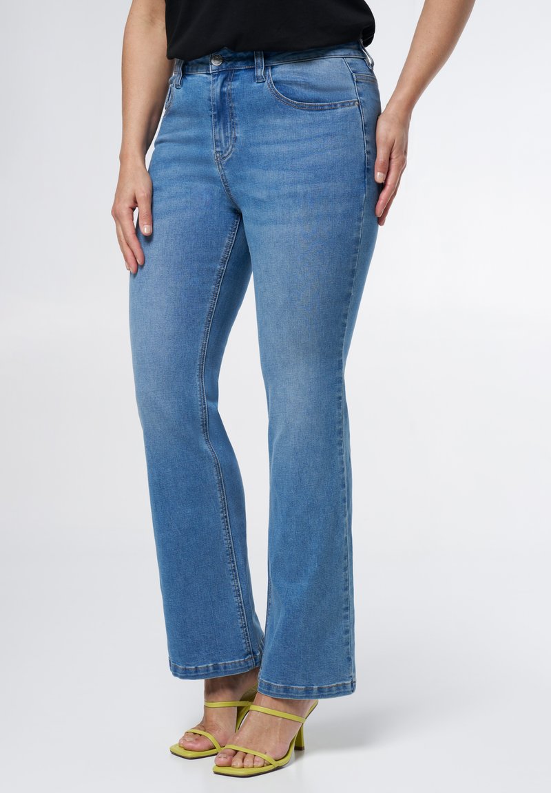 Jean en denim bleu clair évasé avec une taille mi-haute, cinq poches et une texture lisse. Associés à des sandales à talons verts avec des lanières.